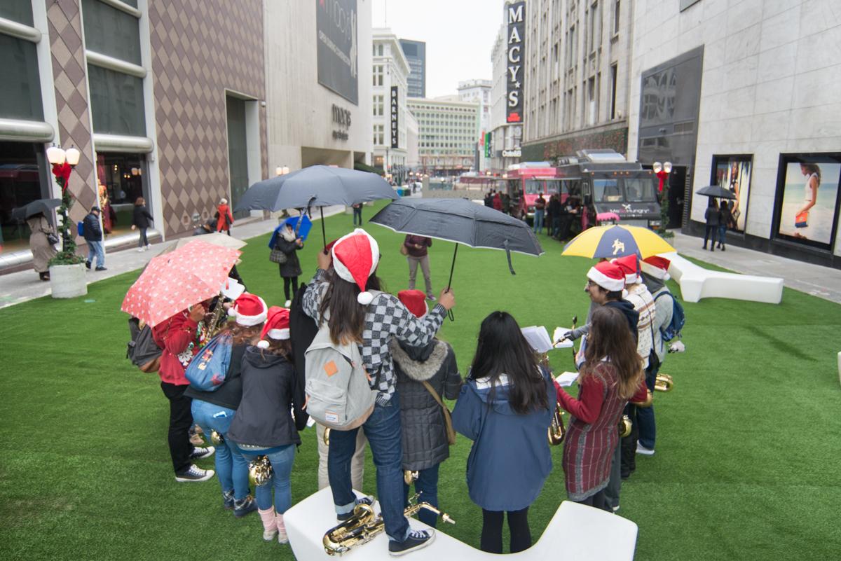 Group of holiday carolers with umbrellas and Santa hats performing on artificial grass in downtown San Francisco plaza.