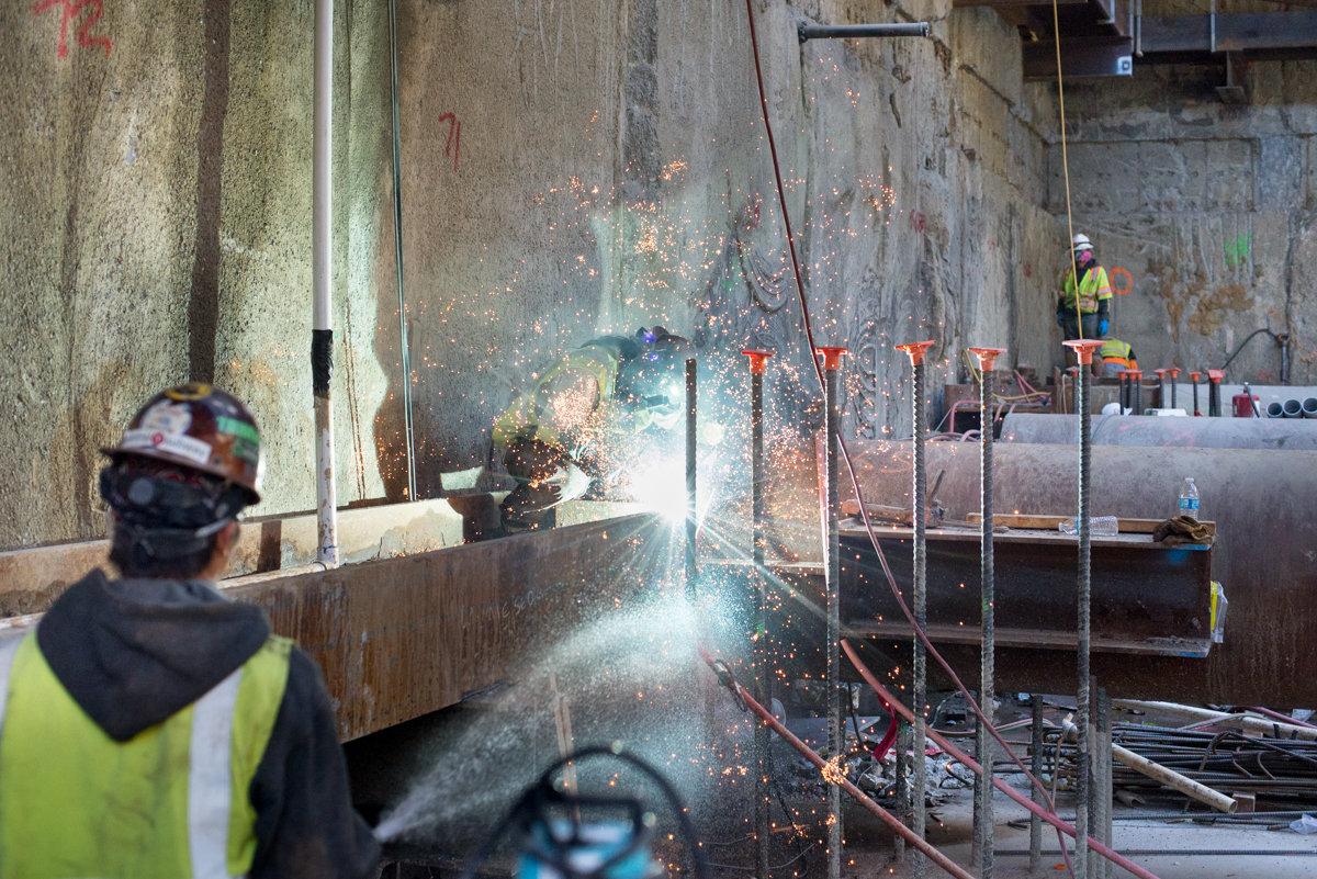 Worker sprays water while cutting steel brackets during underground station construction work.