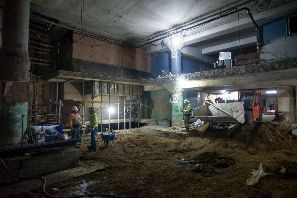 Underground construction site with workers in safety gear, excavation equipment, and structural steel bracing under city streets.