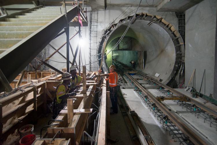 Workers in safety gear construct track foundations inside an underground transit station tunnel with exposed concrete walls and steel reinforcement.
