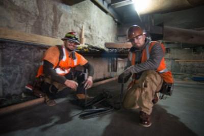 Two workers in orange safety vests and hard hats position rebar sections in an underground transit station construction area.