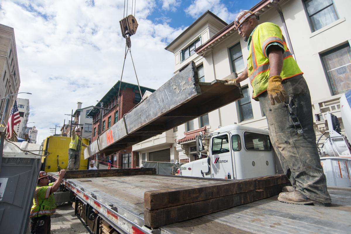 Construction workers guide a steel beam being lifted by crane above an excavation site in an urban area.
