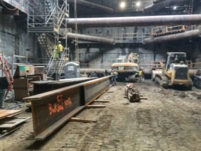 Construction workers and equipment inside underground station headhouse with steel I-beams and structural supports.