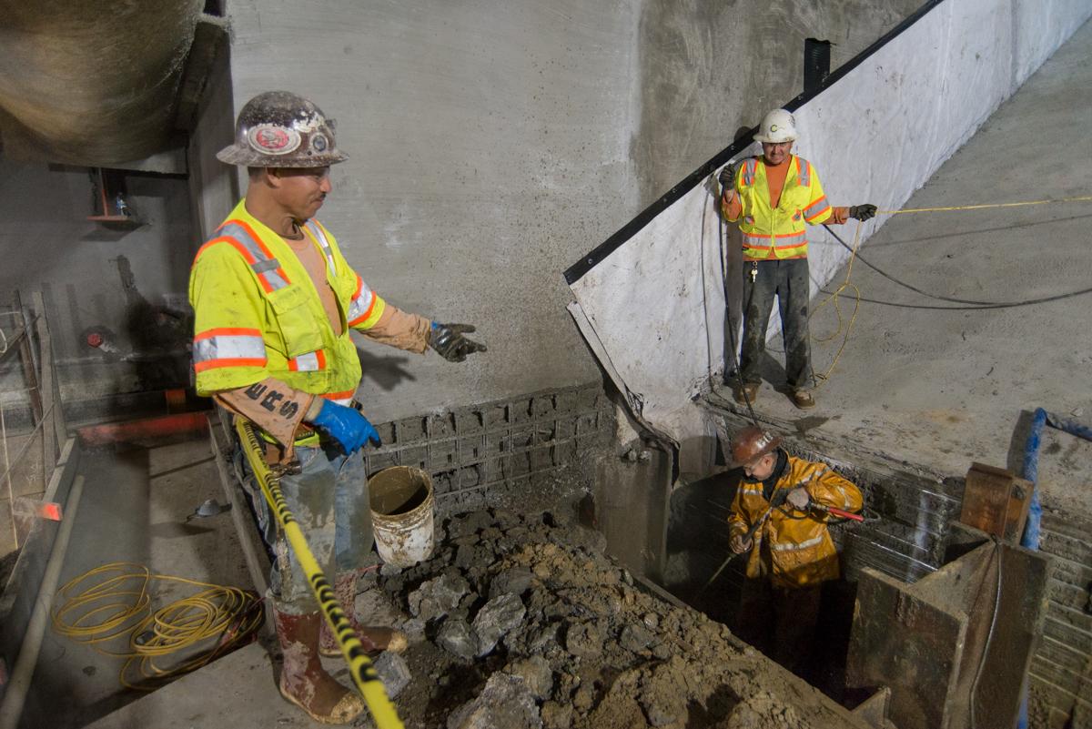 Construction workers spray shotcrete on steel mesh inside underground transit station box.