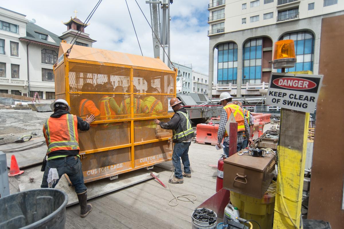 Workers in safety vests operate a yellow construction cage at an urban transit station construction site.