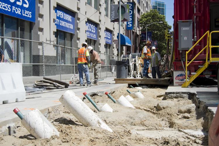 Construction site at urban street with white pipes in sand for ground stabilization, workers in safety vests, and equipment nearby.
