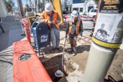 Workers in safety vests examine underground utility work at a street excavation site with exposed pipes and equipment.