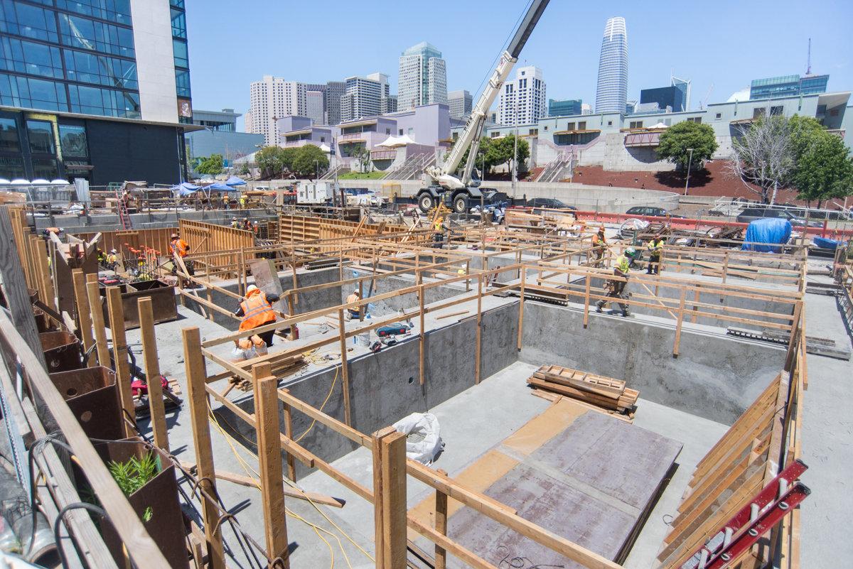Construction site with concrete foundation, wooden framing, and crane near downtown San Francisco skyline.