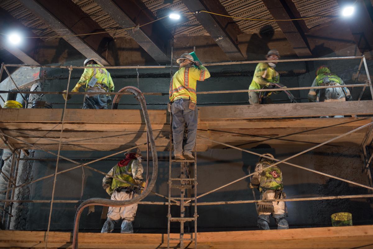 Construction workers in safety vests on scaffolding performing concrete work on interior tunnel walls.