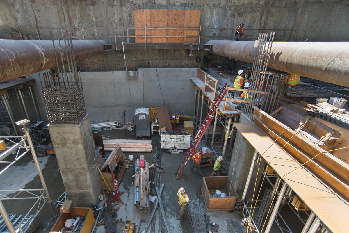 Overhead view of underground transit station construction with workers, scaffolding, and concrete forms on platform level.