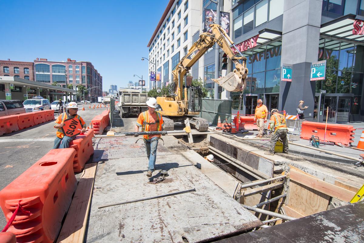 Construction crew operates excavator during water line installation on 4th Street in urban area.