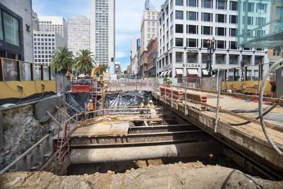 Construction site at Union Square Market Street Station with I-beams supporting station box, downtown San Francisco buildings visible.