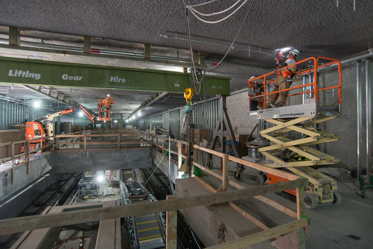Construction workers on scissor lift and boom lift inside underground transit station during utility installation work.