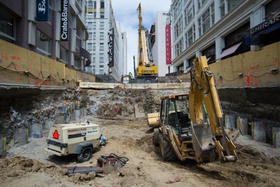 Construction excavation on Stockton Street with exposed beams, heavy equipment, and surrounding buildings.