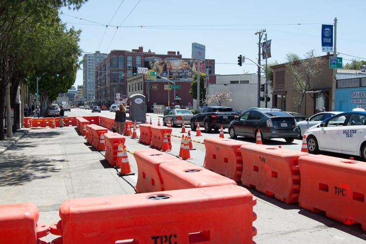 Red plastic barriers and traffic cones block a street work zone on 4th Street, with parked cars and urban buildings visible.