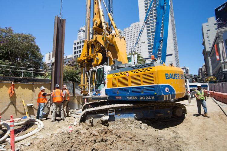 Yellow and blue Bauer excavator on tracked base operates at urban construction site with workers in safety vests nearby.