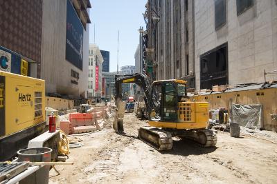 Construction site on urban street with yellow excavator, equipment, and barriers between buildings.