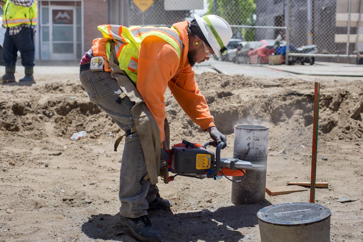 Worker in safety gear cutting a vertical pipe for underground water line valve access.