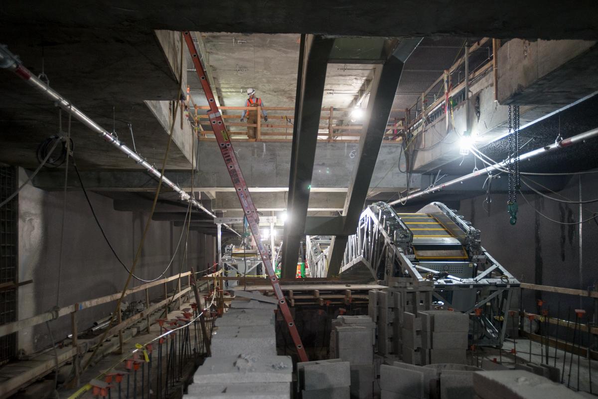 Construction site with concrete pillars, metal stairs, and workers under bright lighting in underground transit station.