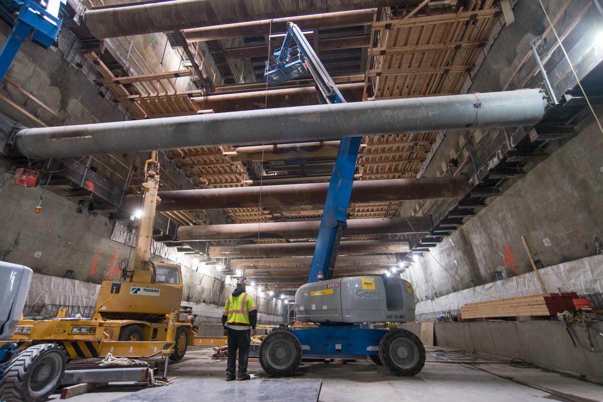 Workers operate boom lifts to install temporary bracing inside an underground station excavation.