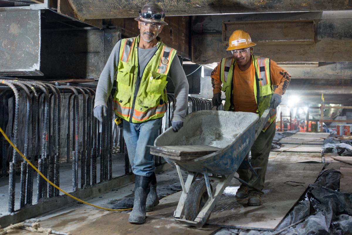 Two construction workers transport materials in a wheelbarrow inside an underground station excavation.