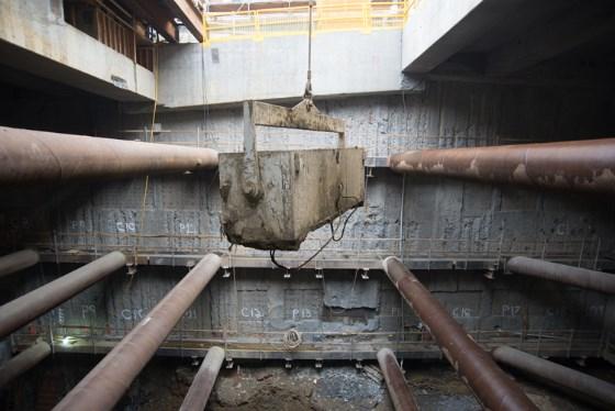 Overhead view of underground transit construction showing steel pipes and an excavation bucket suspended from above.