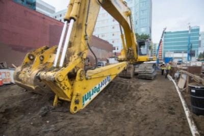 Large yellow excavator with extended arm on construction site near buildings and other equipment.