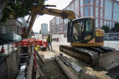Excavator operating in street trench with construction barriers and urban buildings in background.