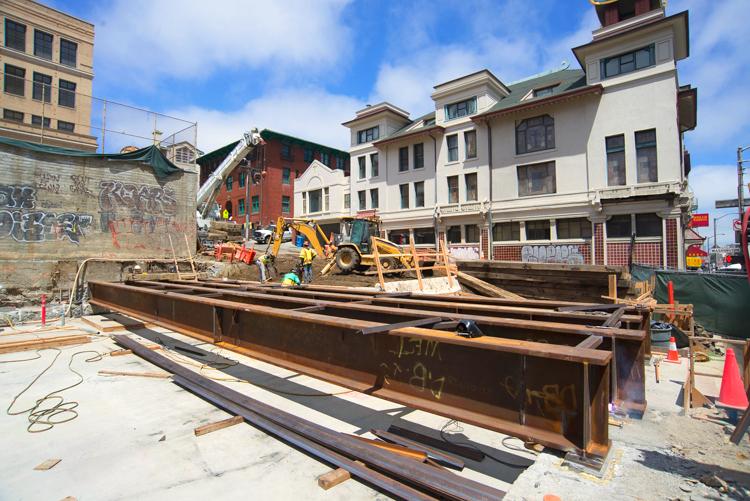 Construction site with large steel beams and heavy equipment near historic buildings in urban area.