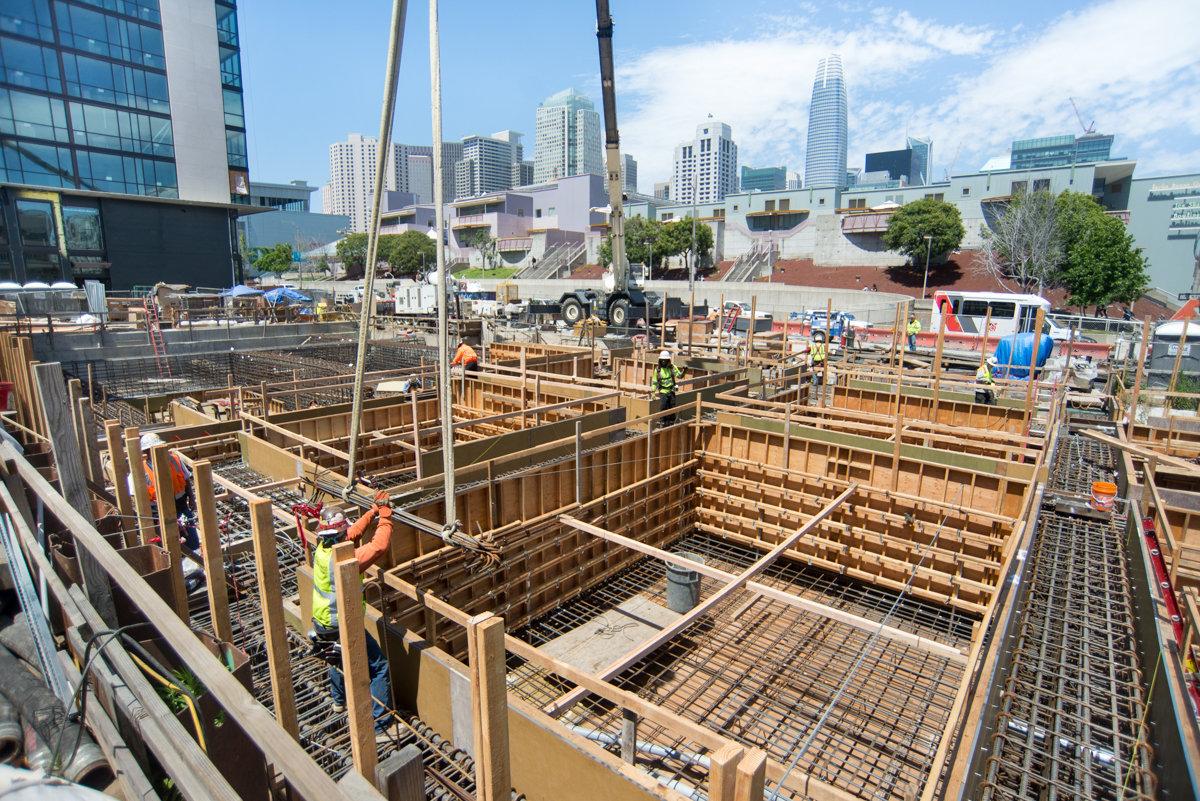 Construction site with wooden shoring, rebar, and crane work on 4th Street with San Francisco skyline in background.