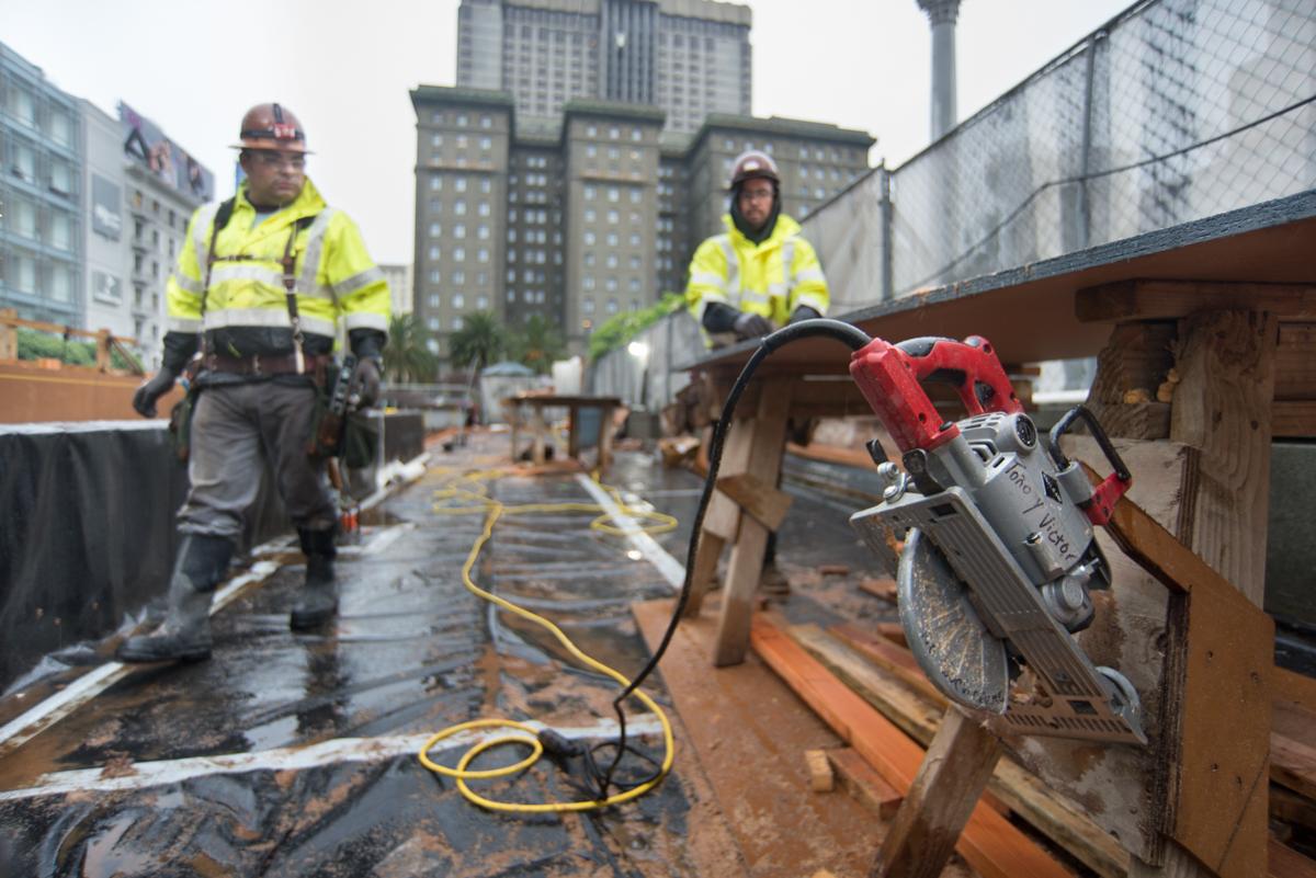 Construction workers in safety vests operate a circular saw on a wet job site with city buildings in the background.