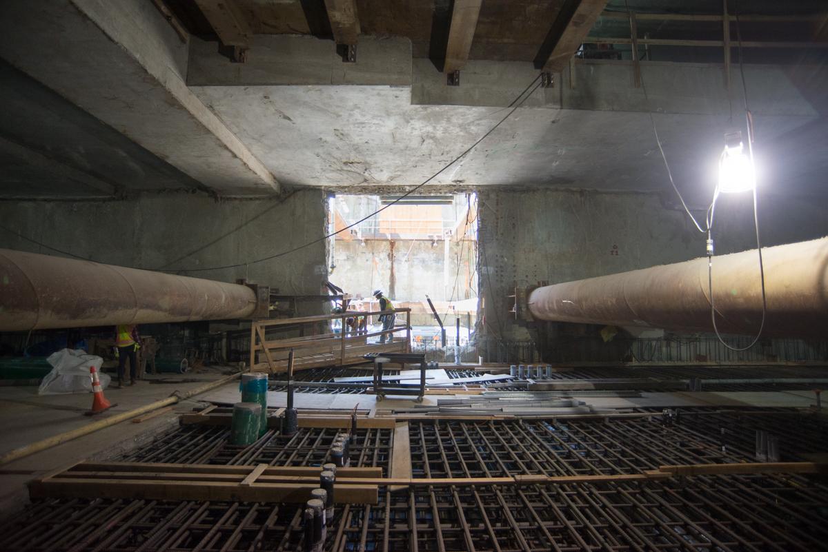 Construction site under concrete bridge with rebar framework and large pipes visible.