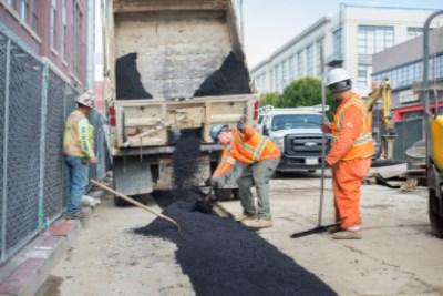 Workers in orange vests unload asphalt from a truck onto a street construction site.