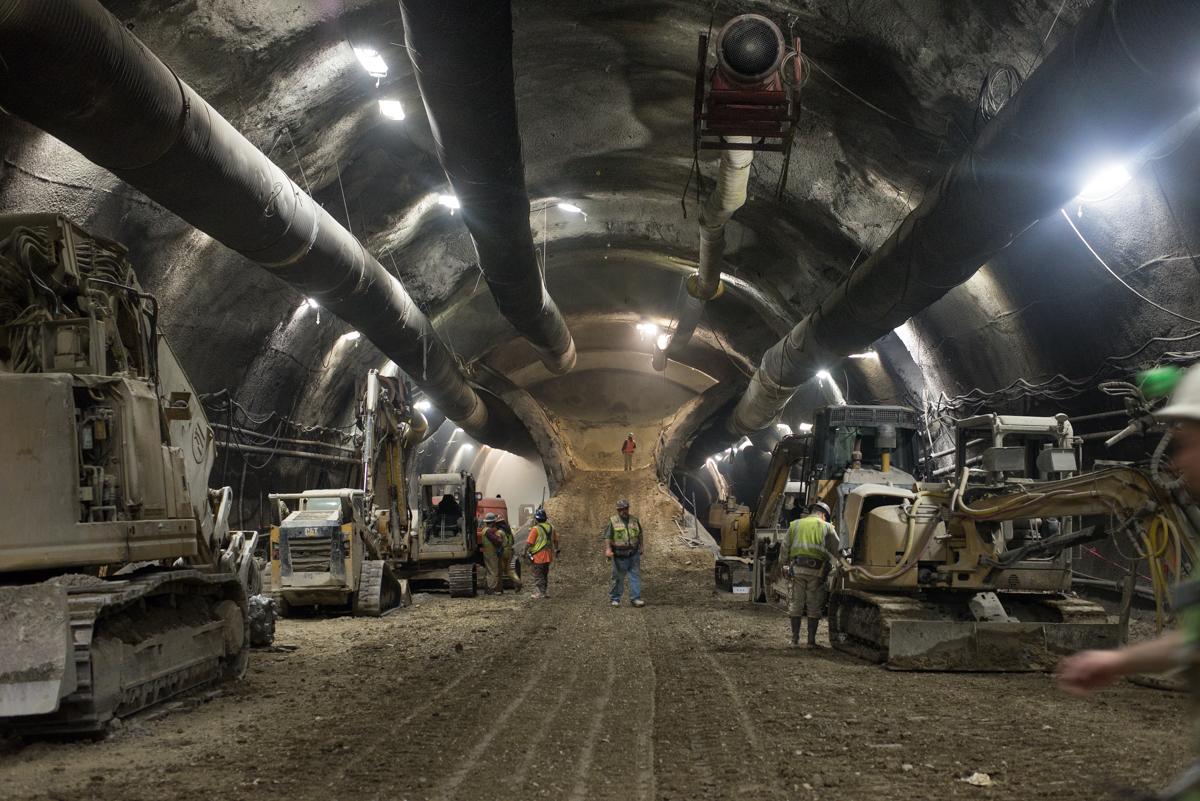 Workers in hard hats inspect a large underground tunnel cavern with heavy equipment and LED lighting.