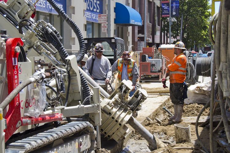 Construction crew operates ground improvement equipment on urban street with buildings and traffic controls visible.