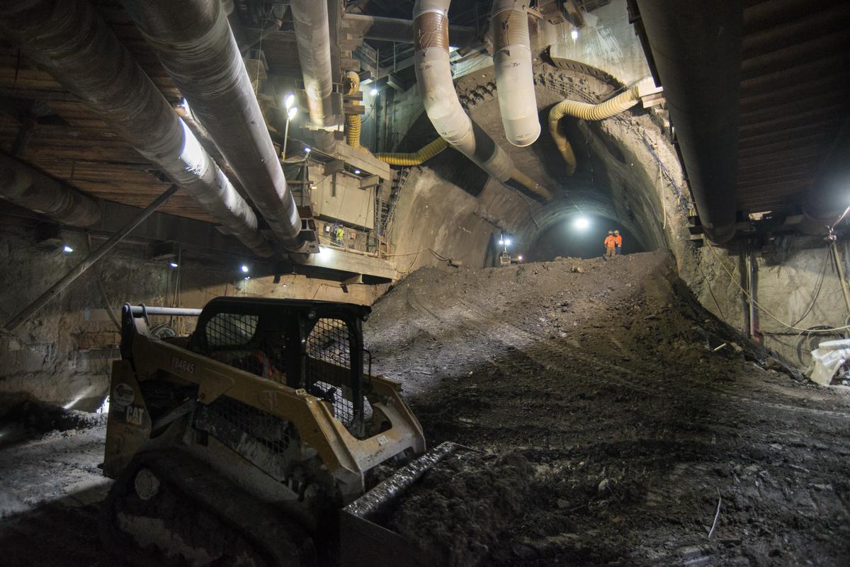 Skid steer loader operating in underground tunnel during Central Subway construction with pipes and lighting overhead.