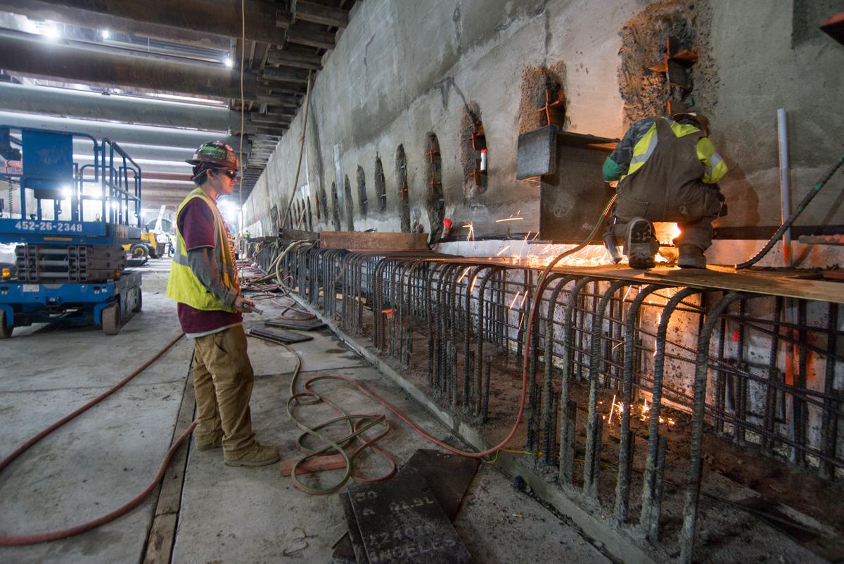 Workers remove temporary steel bracing from a concrete wall during transit infrastructure repair.