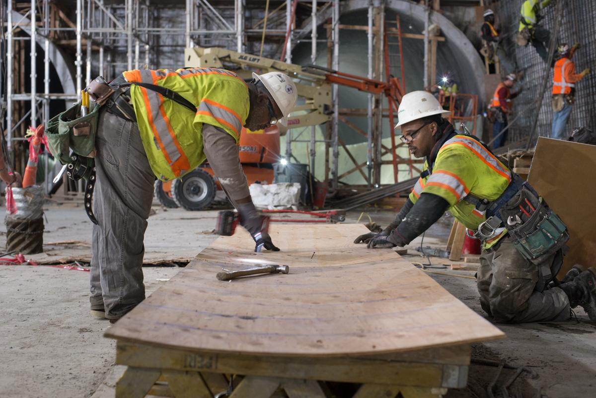 Two construction workers in safety vests and hard hats build a wooden concrete form on a transit station construction site.