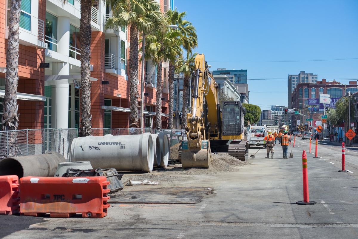 Construction site with yellow excavator, concrete pipes, and workers on urban street with palm trees and red barriers.