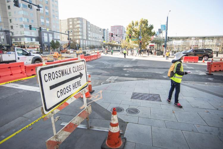 Crosswalk closed sign with construction barriers and worker in safety vest on urban street.