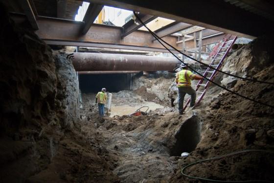 Workers in hard hats excavate underground tunnel with large pipe visible above.
