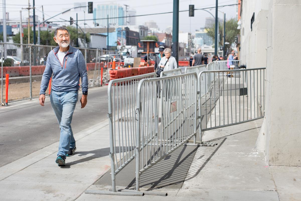 Man jogging past metal barriers on a restored sidewalk section with urban construction site in background.