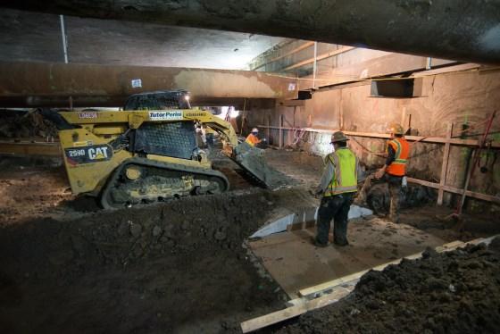 Construction workers and a yellow CAT excavator work on underground station mezzanine floor construction.