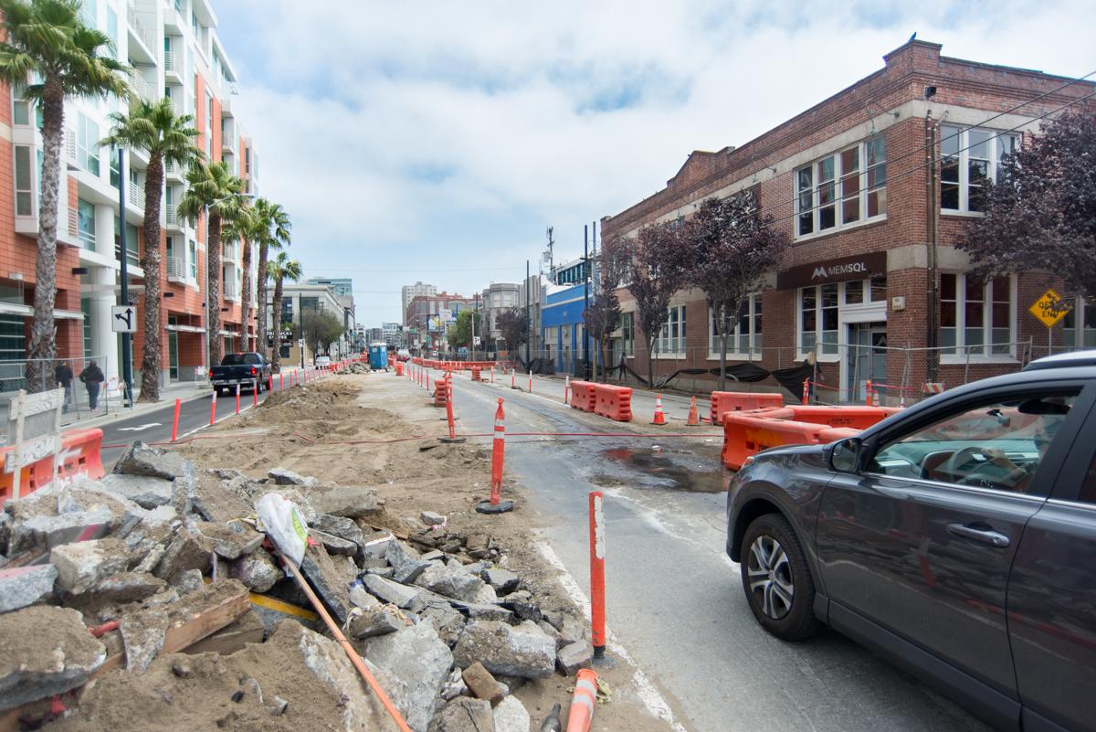 Street construction on 4th Street with traffic barriers, excavated pavement, and surrounding buildings.