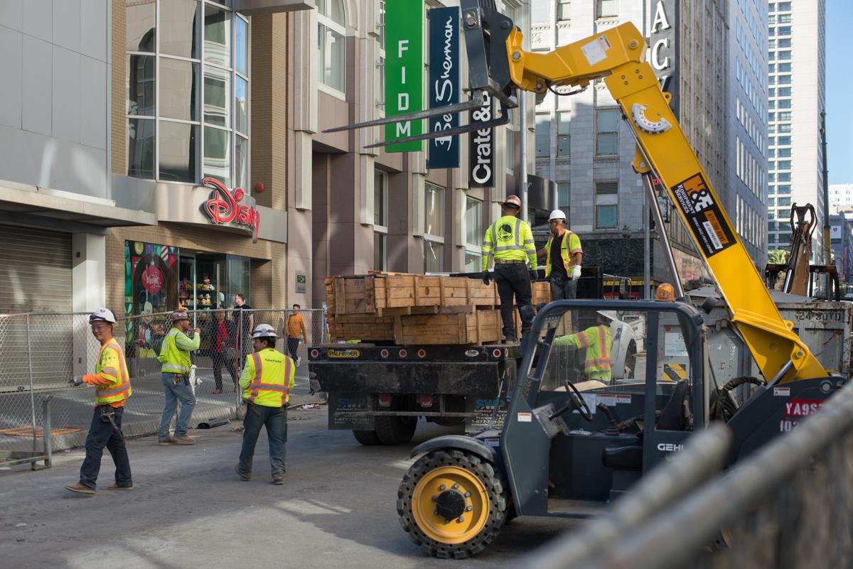 Construction crew unloads wooden crates from truck using yellow crane at Stockton and O'Farrell worksite.