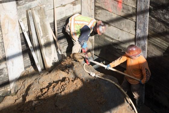 Two workers in safety gear remove soil from inside a subway station access shaft near a shoring wall.
