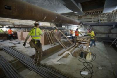 Workers in safety vests install rebar and operate equipment inside an underground transit station excavation.