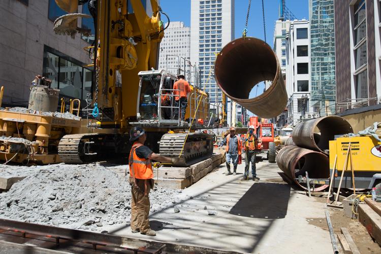 Construction site with heavy equipment, steel pipe casings, and workers in safety vests on an urban street surrounded by tall buildings.