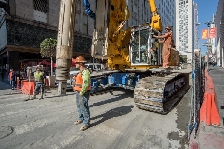 Construction equipment and workers on Stockton Street with orange safety barriers and urban buildings.