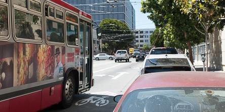 Red transit bus and parked vehicles on city street with buildings in background.
